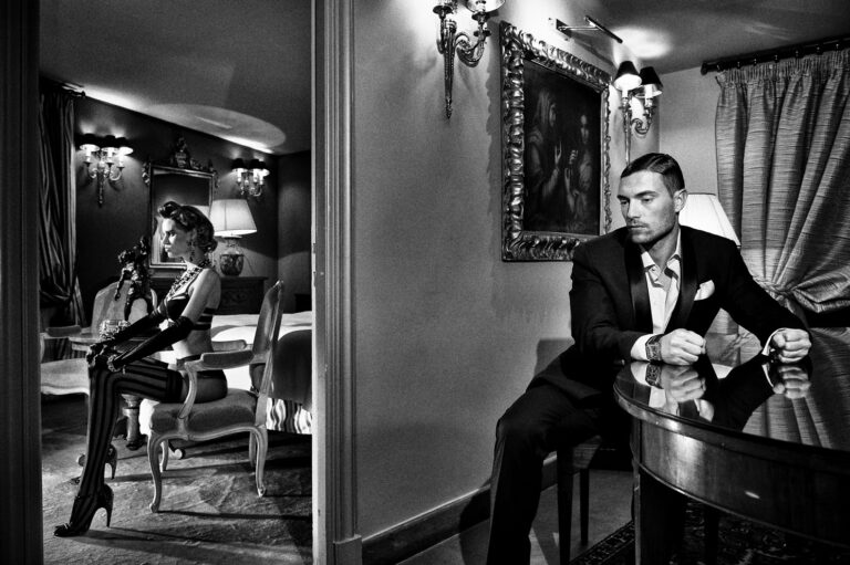 A man in a tuxedo sits alone at a table; a woman in lingerie poses in the adjoining room. Both are framed in a split composition, lit with stark, noir-style lighting.